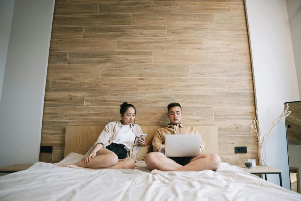 A young couple sitting on a bed using a laptop and a phone in a modern bedroom setting.